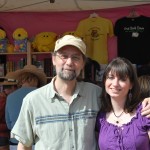 Man wearing a tan hat and green shirt has his arm over the shoulders of a white woman with dark hair wearing a purple shirt. In the background is a booth with books and t-shirts.