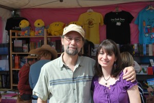 Man wearing a tan hat and green shirt has his arm over the shoulders of a white woman with dark hair wearing a purple shirt. In the background is a booth with books and t-shirts.