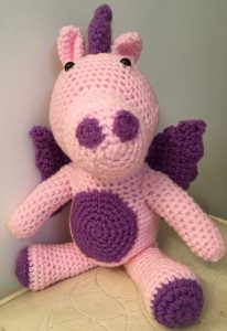 Crocheted dragon. Main body is pink, with dark purple accents - nose, belly, wings, and spines.