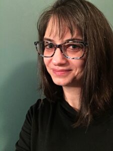 Female leaning against a green wall. She has dark hair, and is wearing a pair of blue and brown glasses. She is smiling slightly.