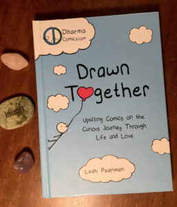 Book cover. It is mostly blue, with line art drawings of clouds that say Dharma Comics and the author's name, Leah Pearlman. The title is Drawn Together, and the O is replaced by a heart balloon. The book is on a wood desktip, with three stones on its left side: pink quartz, green engraved with the word laugh, and purple amethyst.
