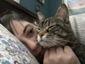 Photo is framed slightly askew. Left side is a pillow with small blue and purple flowers. Right side, in the foreground, is a tabby cat with her eyes closed. Her chin rests on a closed, white-skinned hand. She partially blocks a white female face. You can see her dark bangs, left eye, nose, and a little bit of her lips. She looks very tired. In the top of the photo is a teal-colored wall, with colorful art that’s hard to see or describe.