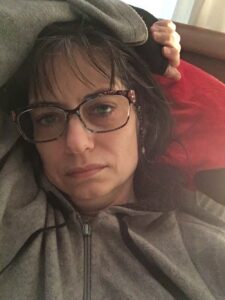 White woman wearing grey hoodie that is zipped up to her neck. Her left arm is flung over her head. She has dark hair and is wearing multi-colored glasses with a beaded glasses chain. She has green eyes. Her expression is neutral. She is lying on a red-and-black pillow.