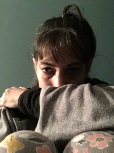 Dark-haired, olive-skinned female sits against a greenish wall. Her dark hair is pulled back in a messy ponytail. She is wearing a black long-sleeved t-shirt under a grey hoodie. Her left arm is across her body, resting on her pajama-clad knees, and obscuring the bottom part of her face. The pajamas are patterned grey, yellow, and pink. The left side of her face is in darkness, and the right side is illuminated.
