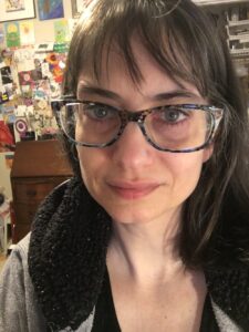 Selfie of girl wearing grey and black hoodie. She has dark hair and blue glasses. The background is colorful - lots of cards and notes on a bulletin board.
