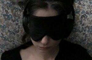 Grainy closeup of light-skinned female face lying on a patterned pillow with blueish, yellowish, and purplish flowers. The eyes are covered by a black sleep mask. The person is wearing a pair of black headphones. She also wears a black shirt that is askew on her shoulders.