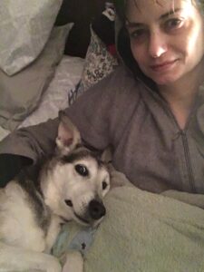 Medium-sized dog with white fur on her face, legs, and belly, and black fur on her head, ears and back, lies on her back next to a white female in bed. The dog looks like she is smiling. The female is faintly smiling, and wearing a grey hoodie. She lies on several pillows and is covered by a green blanket.