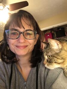 White female with dark, messy hair wearing blue glasses and a grey zipped hoodie on the left. She is grinning. On the right is the profile of a tabby cat’s head. The photo is skewed, so the ceiling fan is above her in the top left corner, and there are some boxes in the top of a closet behind them.