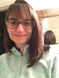 Close-up of white female wearing blue glasses and small stud earrings. Her straight, dark hair goes just past her shoulders on the left side, and is tucked behind her right ear. She is wearing a light turquoise zipped sweatshirt. The back of her head can be seen in the bamboo-framed mirror behind her.