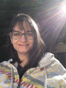 White female with dark hair that goes past her shoulders. She is smiling and wearing blue glasses and a cream-colored hoodie with a pink, blue, and yellow pattern on it. Behind and to her right is a sun flare, so there are light beams around her.