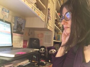 On left side of photo, side view of white female. Her dark hair is down, and she is resting her chin on her right hand. She is wearing a purple shirt. She’s staring at a laptop screen (on the left of the photo), and the light is reflected in her blue glasses. She is sitting at a desk, and there is a mess of books and paper and colorful things around her.