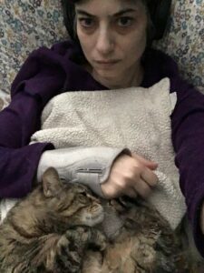 At the top of the photo, the face of white female is laying on a patterned pillow with blueish, yellowish, and purplish flowers. She is wearing black headphones. Her expression shows pain. She is wearing a purple top and a brace on her right wrist. She is covered by a white blanket. The bottom of the photo is a tabby cat curled up with her head pressed against the brace and her feet pressed against the hand. Her right paw is curled under her chin.