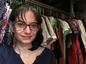 White female wearing blue v-neck top and blue glasses. Her dark hair is messy. She is smiling slightly. She is leaning against a row of colorful clothes hanging in her closet, mostly skirts and tops.