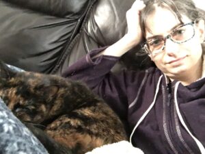 Angled photo of a black couch. On the left is a curled up tortoiseshell kitty on a blue and white blanket. She appears to be sleeping. On the right is a white female wearing patterned glasses and a zipped-up purple hoodie. Her dark hair is messy. She is leaning her head on her right hand; her elbow is resting behind the cat. She looks tired.