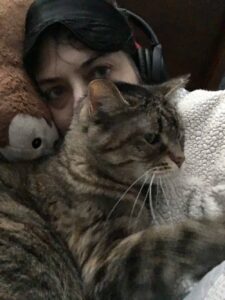 Foreground of photo shows a tabby cat whose head is partially blocking a white female face. The human is wearing black headphones, and her head is on a round, brown pillow. Her eye mask is on her forehead. She is covered by a white blanket.