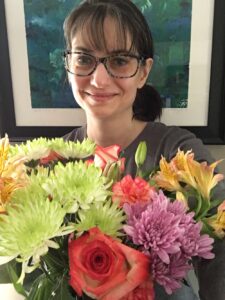 White female with dark hair pulled into a small ponytail on the left side of her head. She is smiling and wearing blue glasses, and holding a bouquet of various pink, yellow, purple, and green flowers in front of her. The background is a black-framed and white-matted abstract painting in blues, greens, and whites.