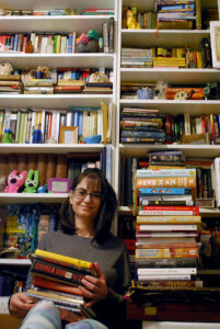 White female sitting in bottom middle of photo. She has dark hair and is wearing blue glasses, a grey shirt, and pink and blue multi-colored patterned leggings. Two very full bookshelves are behind her, and a stack of books is beside her. She is holding a small stack of books.