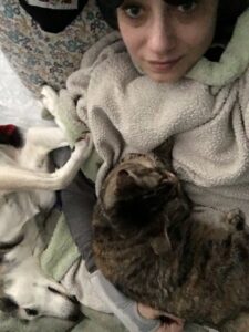 Photo from above shows a white female face. She is on top of several multi-colored pillows, and under a white blanket. A tabby cat sits curled on chest, and her arm is wrapped around the cat. The woman is wearing a dark grey shirt and a light grey hand brace. To their right is a small dog with white fur and black snout, resting on her side. The dog’s head is at the bottom of the photo, and her paws are angled between the cat and the person’s head.