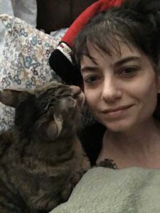 Tabby cat (on the left) with her paw on a white female’s chest. The cat is licking the cheek of the girl (on the right), who is in bed under a green blanket. Her head on top of a red pillow, and a pillow with small blueish, purplish, and yellowish flowers. The girl has dark bangs and is smiling slightly.