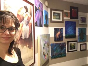 Photo of white female with messy dark hair, wearing blue glasses & black scoop-necked top. She stands next to the wall of paintings listed in text. Descriptions - Kaitlyn: pink flower on a black background; mine: abstract tree on hill with blue sky; Karin: marsh scene with a willow tree draped over the top of the photo; hubs: blue turtle on an orange background; Karin: closeup of red apple; & photo by Kaitlyn: a pink flower. Big square painting: the circle mandala is made up of a series of colorful circles in purple, white & black, orange, reddish, green, white & black yin-yang, yellow tennis ball, red target logo, blue earth, orange & brown, black, and green. Second row: pink crane on blue background; by me: abstract blue sky with green grass on the bottom, & brown turtle on blue background. The bottom row: by me: abstract reddish & bluish landscape; the winning photograph: mostly greens; tiny tiny paintings: a number of little rectangles in in different colors.