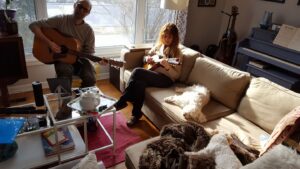 Photo of two people in a living room. The male is holding a guitar, and the red-haired female is playing a mandolin. Two white dogs are asleep on the couches next to her. A black piano is in the far right of the photo.