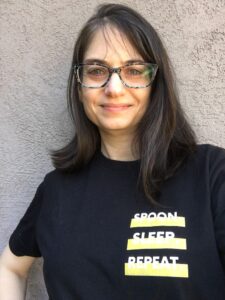 White woman with blue glasses leaning against a brown stucco wall. Her dark hair is down, and she is smiling. She is wearing a black t-shirt with the words Spoon Sleep Repeat on it in white.
