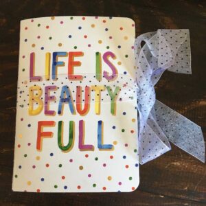 A full-looking notebook tied shut with a sheer organza ribbon with black dots. The cover is colorful and says Life Is Beauty Full.
